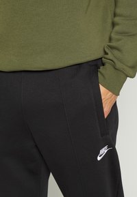 Černé tepláky s bočními kapsami a bílým logem Nike; spárované se olivově zeleným tričkem s dlouhým rukávem vyrobeným z jemného materiálu.