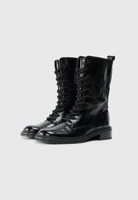 Bottines en cuir noir à lacets avec une finition brillante, dotées d'un bout rond, de lacets noirs et d'un petit talon. Accents texturés le long des coutures.