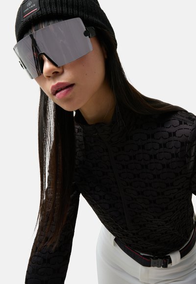 Μαύρο μπλουζάκι με φερμουάρ και μακρύ μανίκι με διακριτικό σχέδιο, συνοδευόμενο από μεγάλα γκρίζα γυαλιά ηλίου και μαύρο πλεκτό σκουφάκι. Λευκό παντελόνι με ζώνη.