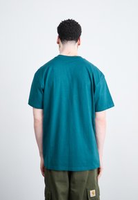 Teal cotton T-shirt with a round neckline and short sleeves, featuring a straight cut and smooth texture, worn with olive cargo pants.