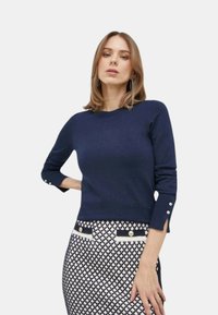 Maglione in maglia blu navy con maniche a tre quarti, caratterizzato da tagli ai polsini e dettagli con bottoni. Abbinato a una gonna a fantasia con dettagli a contrasto.