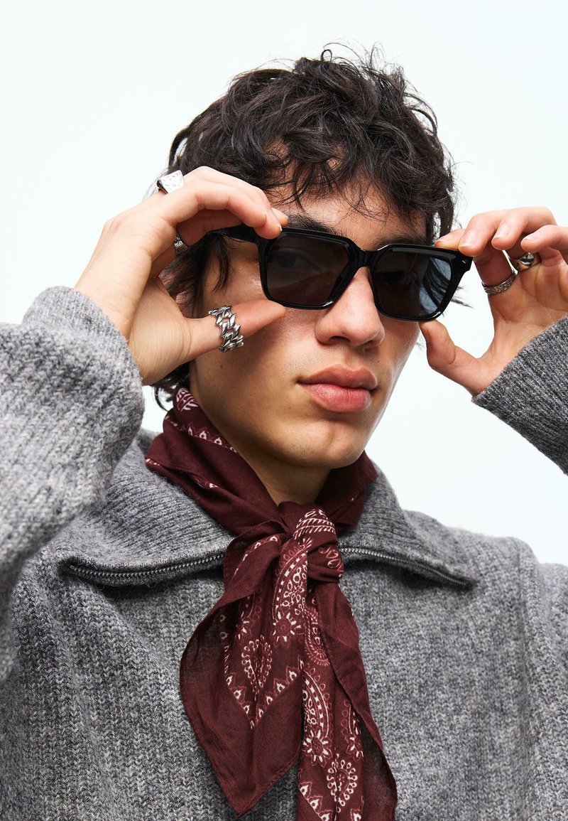 Jeune personne ajustant des lunettes de soleil carrées noires, portant un pull tricoté gris et une écharpe à motifs bordeaux autour du cou, avec des cheveux bouclés et foncés.