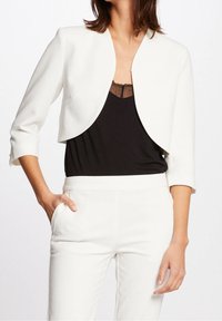 Kvinna iklädd en vit croppad kavaj med trekvartsärmar över en svart topp med spetsdetaljer och matchande vita byxor, med en hand i fickan.