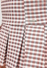 Jupe plissée à carreaux rose, blanc et rouge foncé avec une petite étiquette argentée de la marque "GUESS" sur la ceinture.