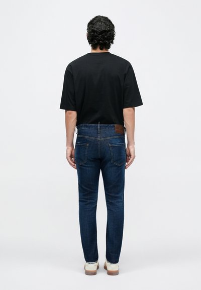 Fekete rövid ujjú póló sötétkék farmernadrággal párosítva. A farmernadrág barna bőr címkével rendelkezik a derékrészen, egyenes szárú szabással.