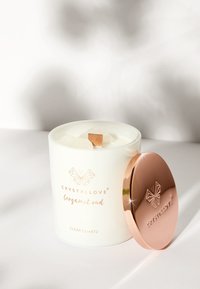 Candela bianca con uno stoppino in legno, contenuta in un contenitore cilindrico. Il coperchio metallico dorato presenta un logo. L'etichetta riporta "CRYSTALLOVE bergamot oud."