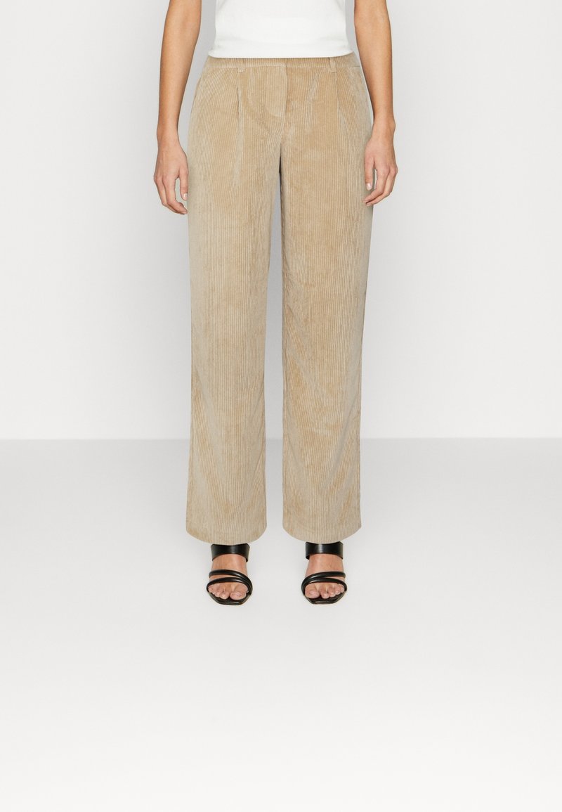 Pantalon beige large en velours côtelé porté avec des talons noirs à brides ouverts et un haut blanc, debout devant un fond clair uni.