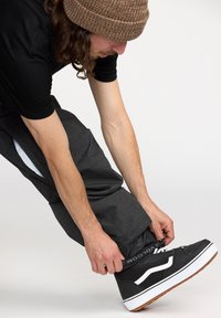 Czarne buty do skate z białymi akcentami, związane i zapięte. Osoba nosi ciemnoszare spodnie z boczną kieszenią na zamek błyskawiczny i siateczkowym wykończeniem.