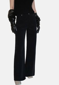 Pantalons larges noirs en tissu texturé, avec une taille haute et des boutons avant en métal argenté. Accompagnés de longs gants noirs.