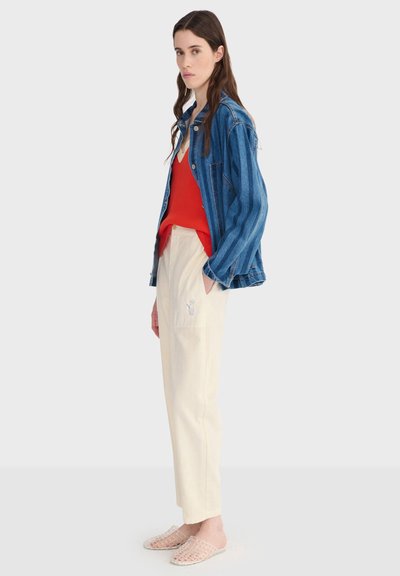 Jeune femme portant une veste en denim bleu, un haut rouge, un pantalon couleur crème avec un petit logo brodé, et des chaussures transparentes tissées sans lacets.