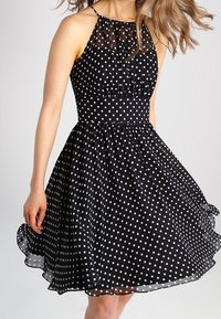 Schwarzes Kleid mit Pünktchenmuster, enganliegendem Oberteil und fließendem Rock, mit einem transparenten Überzug am Ausschnitt und Halterneck-Trägern.
