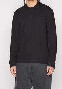 Svart långärmad polo skjorta tillverkad av texturerat tyg med en ribbad design, som har en knappslå med tre knappar och en broderad logotyp på bröstet.