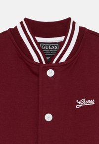 Maroon varšavska jakna s bijelim prugastim ovratnikom, prednjim kopčanjem na dugmad i izvezenim bijelim logom "Guess" na lijevoj busti. Mekana tekstura tkanine.