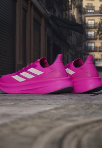 Lyserøde Adidas løbesko med hvide striber, der hviler på en byvej med bygninger i baggrunden.
