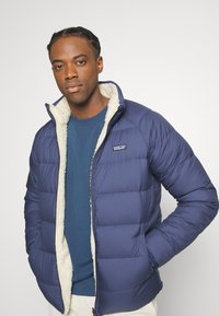 Veste matelassée marine avec un design matelassé, doublure en fausse fourrure crème, fermeture éclair, et logo Patagonia sur la poitrine. Portée sur un pull bleu.
