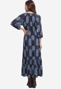 Kobieta z długimi włosami, ubrana w długą sukienkę w patchworkowym stylu w granatowym kolorze w wzór paisley oraz brązowe botki, stoi z jedną ręką na plecach.