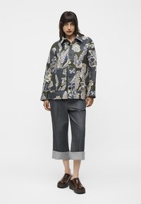 Mujer con chaqueta de estampado floral oscuro, pantalones grises de pierna ancha con puños y zapatos marrones de plataforma sin cordones, de pie frente a un fondo simple.