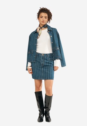 Kvinde iført blå denimjakke med striber over hvid top, matchende denimnederdel, sorte knæhøje støvler og mønstret halstørklæde.