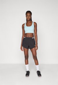 Ljusblå sport-bh med breda axelremmar, i kombination med svarta löparshorts. Modellen har på sig vita strumpor och svarta sportskor med rosa detaljer.