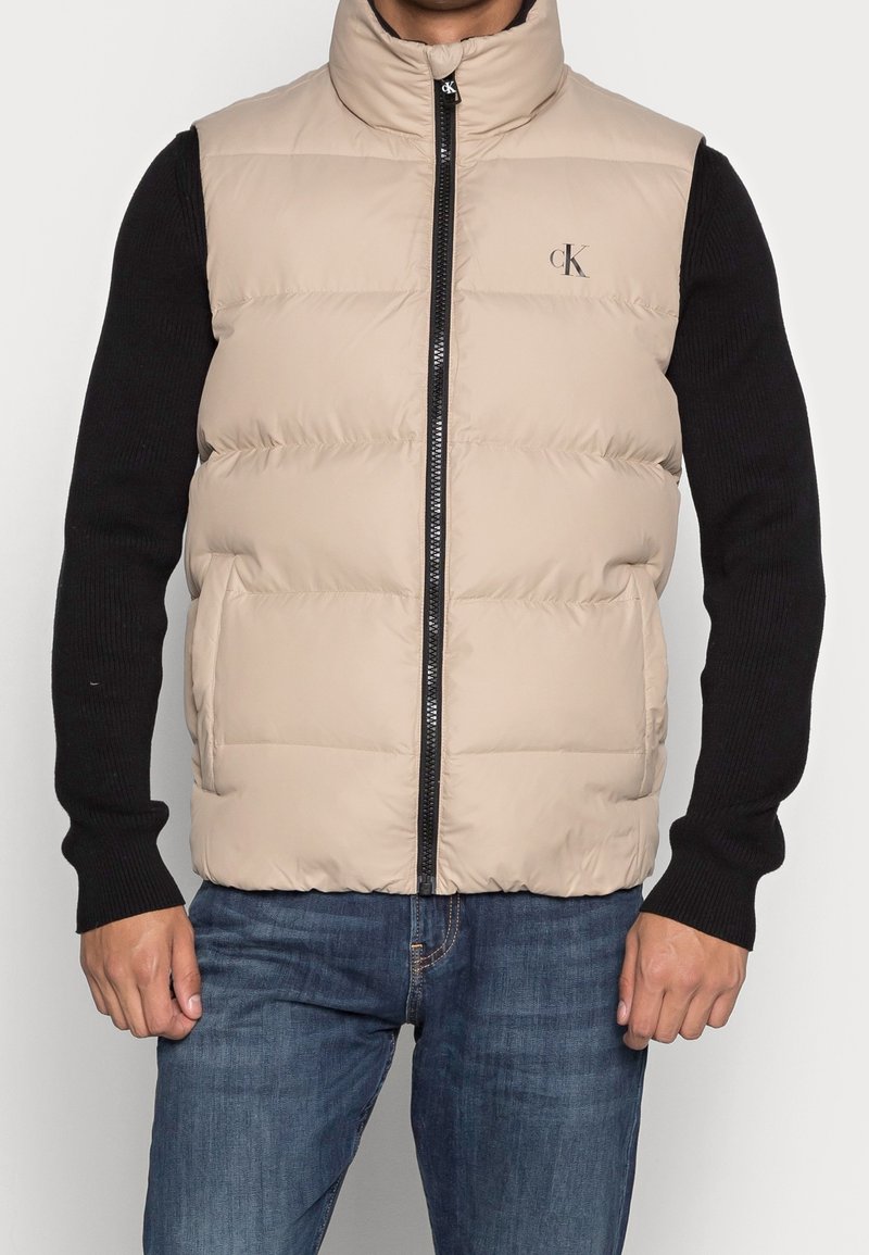 Beige pufferväst med hög krage, frontdragkedja och svart logotyp på vänster bröst. Texturerad yta och sidofickor. Svart långärmad tröja under.