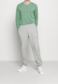 Zielona koszulka z długim rękawem i szare spodnie dresowe z elastycznymi mankietami, z małym białym logo na piersi i spodniach. Białe trampki.