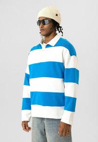 Polo à manches longues rayé bleu et blanc avec col, associé à un jean en denim clair et un bonnet en maille beige. Comprend des lunettes de soleil foncées.