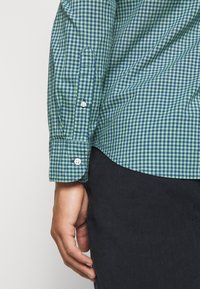 Camicia a quadri verde e blu navy con polsini abbottonati e orlo inclinato. Il tessuto sembra leggero e liscio. Mostra parte di una mano.