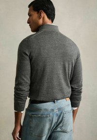 Polo gris à manches longues avec un col haut et des poignets côtelés, porté par-dessus une chemise blanche, associé à un jean en denim bleu clair.