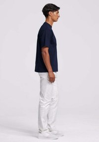 Donkerblauwe katoenen t-shirt met korte mouwen, rechte pasvorm. Gecombineerd met witte broeken en grijze sneakers, van opzij getoond.