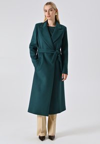 Lang, teal wollen jas met een ceintuur in de taille, reverskraag en zijzakken; gedragen over beige pofbroek en puntige schoenen.