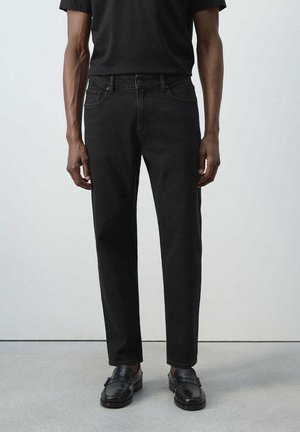 Hombre con vaqueros negros rectos, camisa negra y mocasines de cuero negro, de pie sobre suelo gris frente a pared blanca.