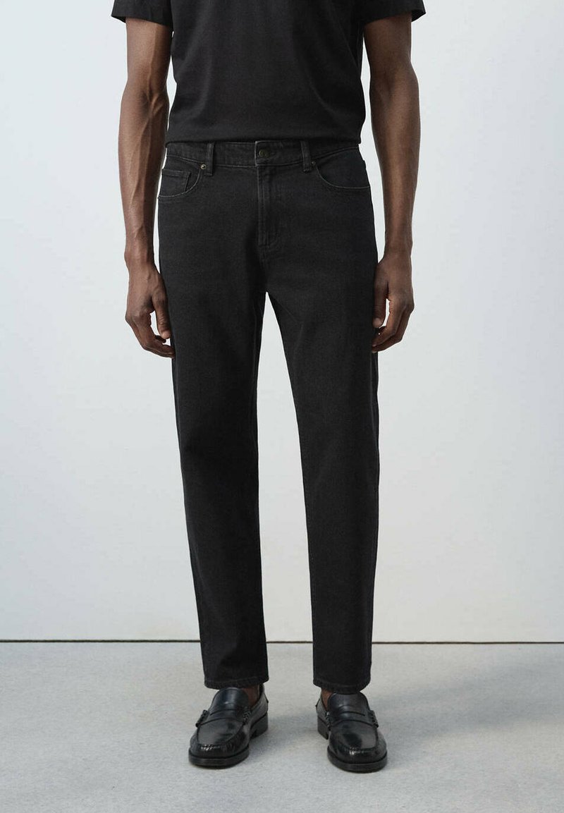 Homme portant un jean noir droit, une chemise noire et des mocassins en cuir noir, debout sur un sol gris contre un mur blanc.