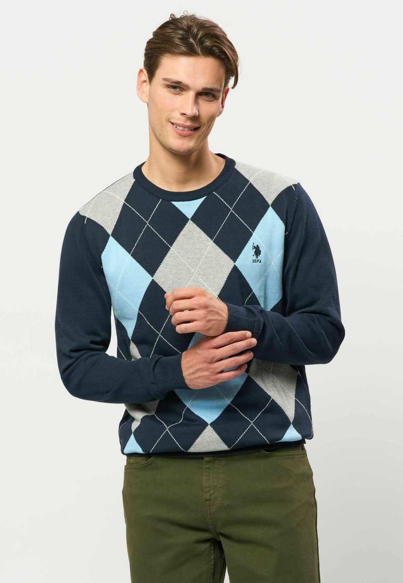 Jeune homme souriant portant un pull à losanges bleu marine, bleu clair et gris avec un pantalon vert olive sur un fond uni.