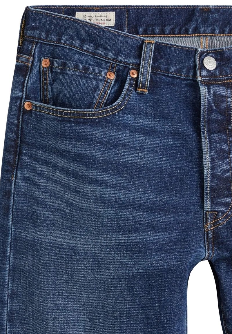Jean en denim bleu foncé en gros plan montrant les poches avant, les rivets en cuivre, les passants de ceinture, la fermeture par bouton et une étiquette en tissu blanc à l'intérieur de la ceinture.