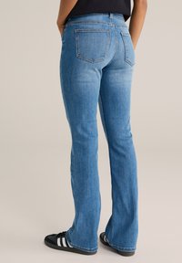 Kék farmer trapézszárú nadrág középmagas derékkal, hátsó zsebekkel és puha anyaggal, fekete cipőkkel, fehér csíkokkal párosítva.