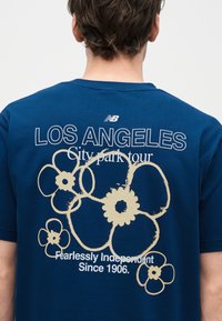 Άτομο που φοράει μπλε μπλουζάκι με μπεζ λουλουδένια σχέδια και κείμενο στην πλάτη που γράφει "LOS ANGELES City park tour Fearlessly Independent Since 1906".