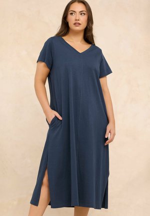 Kobieta ubrana w granatową sukienkę midi z krótkim rękawem i dekoltem w serek, z bocznymi kieszeniami i rozcięciami, stojąca na beżowym tle.