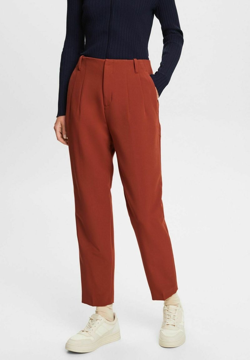 Personne portant un pantalon fuselé couleur rouille, un top noir côtelé à manches longues et des baskets crème, debout avec une main dans la poche.