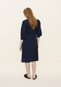 Navy-blaues Kleid mit Puffärmel und gerüschtem Rücken, knielang, aus einem leichten Stoff gefertigt. Getragen mit dunkelbraunen Slippern.