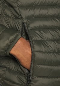 Olivgrüne Stepp-Daunenjacke mit horizontalem Steppmuster, ausgestattet mit einer Reißverschlusstasche und elastischen Bündchen. Glattes, leichtes und isoliertes Material.