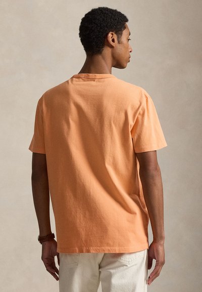 Άνδρας που φοράει απλό ροδακινί κοντομάνικο T-shirt και ανοιχτό μπεζ παντελόνι, γυρισμένος προς τα πίσω με φόντο ουδέτερο μπεζ.