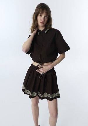 Femme portant une chemise noire à manches courtes boutonnée et une jupe assortie avec une broderie beige paisley près de l'ourlet, debout devant un fond clair uni.