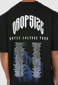 Sort bomulds T-shirt med teksten "DROPSIZE HUSTLE CULTURE TOUR" i fed hvid skrift, med en liste over byer trykt nedenfor.