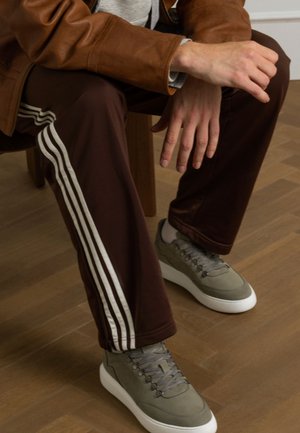 Bruine sportbroek met witte strepen en een grijze casual schoen met een gestructureerde bovenkant en een witte zool, zittend op een houten vloer.