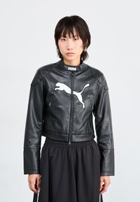 Veste en cuir noir, raccourcie, avec col montant, présentant un grand logo Puma blanc sur la poitrine et deux poches zippées à l'avant.