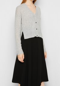 Grauer Strickcardigan mit V-Ausschnitt, langen Ärmeln und schwarzen Knöpfen. Kürzerer Schnitt mit geripptem Saum. Über einem schwarzen Rock getragen.