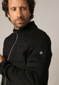 Zwarte vest met een rits aan de voorkant, gewatteerde panelen aan de voorkant, geribbelde kraag en gestructureerde mouwen. Bevat een cirkelvormig logo op de linker mouw.