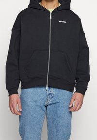 Homme portant un sweat à capuche noir zippé avec le texte "WRSTBHVR" sur la poitrine et un jean bleu, debout devant un fond uni.