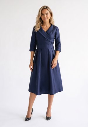 Femme debout portant une robe portefeuille bleu marine avec manches trois-quarts et des talons hauts noirs sur un fond blanc uni.