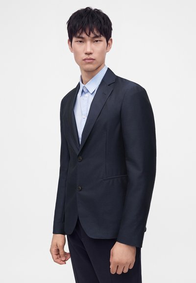 Jeune homme portant une veste de costume bleu marine foncé sur une chemise bleu clair boutonnée, debout devant un fond blanc uni.